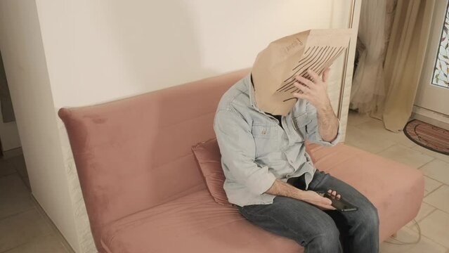 A man wearing a breadbag face, sitting on the sofa at home, looking at his smartphone and feeling ashamed.

