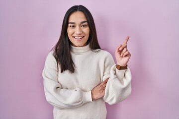 Young south asian woman standing over pink background with a big smile on face, pointing with hand finger to the side looking at the camera.