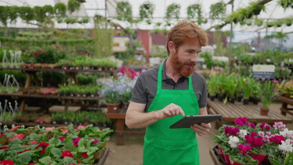 Young man using tablet to browse inventory walking inside Flower Shop. A male employee wearing green apron holding inspecting store with modern technology