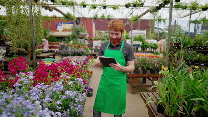 Smiling Red-Haired Employee with Tablet in Flower Shop. Young Man in Apron Leveraging Modern Tech in Local Business