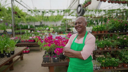 Fototapeta na wymiar Portrait of an African American older woman posing for camera with arms crossed standing inside Local Flower Shop. A black senior female employee wearing green apron