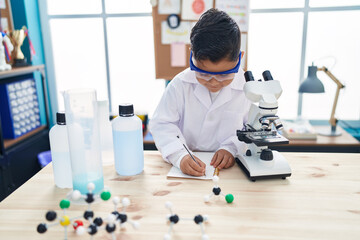 Adorable hispanic boy student using microscope writing notes at laboratory classroom