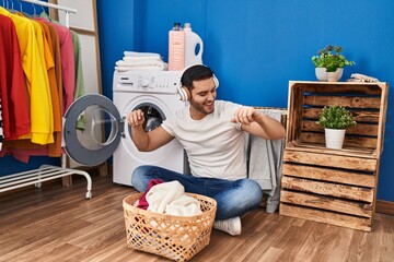 Young hispanic man dancing and listening to music washing clothes at laundry room