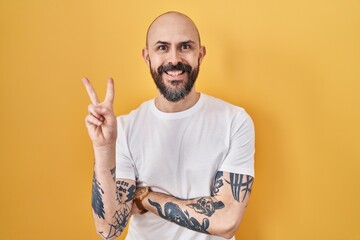 Young hispanic man with tattoos standing over yellow background smiling with happy face winking at...