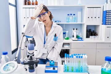 Young hispanic woman working at scientist laboratory surprised with hand on head for mistake, remember error. forgot, bad memory concept.