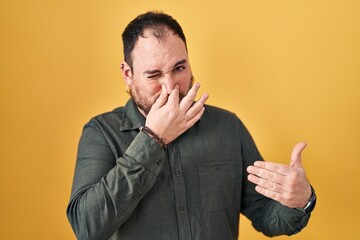 Plus size hispanic man with beard standing over yellow background smelling something stinky and disgusting, intolerable smell, holding breath with fingers on nose. bad smell