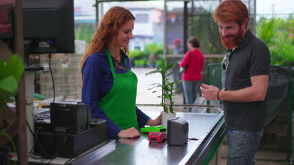 Male Customer Purchasing Plant with contactless payment cellphone at Horticulture Store Checkout. Female employee Assisting Client at Local Business