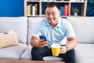 Young chinese man using smartphone drinking coffee at home