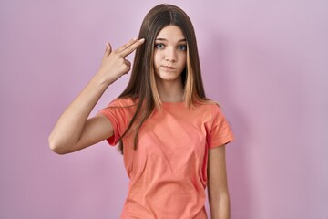 Teenager girl standing over pink background shooting and killing oneself pointing hand and fingers to head like gun, suicide gesture.