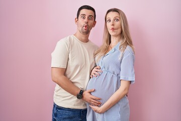 Fototapeta premium Young couple expecting a baby standing over pink background making fish face with lips, crazy and comical gesture. funny expression.