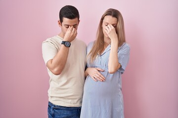 Young couple expecting a baby standing over pink background tired rubbing nose and eyes feeling fatigue and headache. stress and frustration concept.