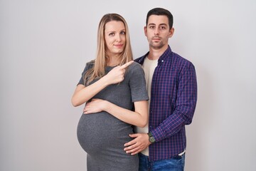 Young couple expecting a baby standing over white background pointing with hand finger to the side showing advertisement, serious and calm face