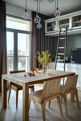 Wooden table with branches in the vase, apples, glass and  four chairs in the modern kitchen. There is white lockers and shelves with accessories, dark ladder, hanging lamps, glass door with a window.