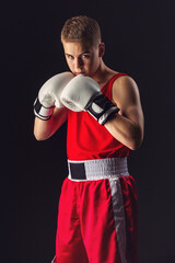 Fototapeta na wymiar Young handsome boxer sportsman in red boxer suit and white gloves standing on black backgound. Copy space.