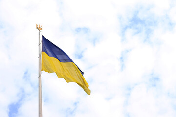 The flag and coat of arms of Ukraine on a flagpole against a cloudy sky. The flag of Ukraine develops in the wind