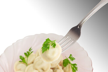 Cooked meat dumpling with a parsley twig on fork on a background of pink glass dish with pelmeni on...