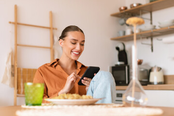Excited lady using smartphone in kitchen while having dinner, sitting at table and texting on...
