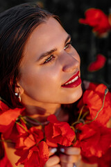 Naklejka premium Beautiful young woman with brown hair in red long dress in field with poppies in evening at sunset and holds poppy in hand, Summer countryside nature flowers, Female relaxing, Rural simple life