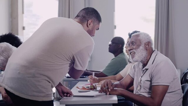 professor Brasileiro na mesa de um aluno idoso explicando a licao de matematica em um curso para adultos e idosos no Brasil