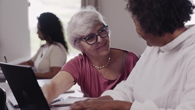 mulher idosa Brasileira ajudando a colaga mais jovem com dificuldade para usar o computador em uma turma para alunos senior no Brasil