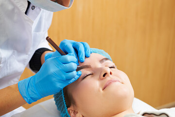 Cosmetician making permanent eyebrow make up for beautiful girl. Eyebrow tattoo. Close up.