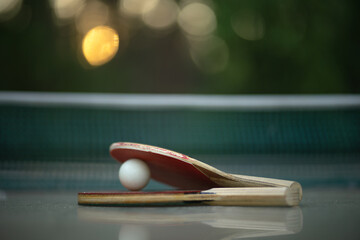 Ping pong rackets and a white ball lie on the table in the rays of the setting sun.Outdoor games in...