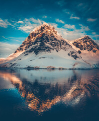 Fototapeta na wymiar Antarctic landscape with snow covered mountains reflected in ocean water. Sunset warm light on the mountain peak, blue cloudy sky in the background. Beautiful nature landscape. Travel background.