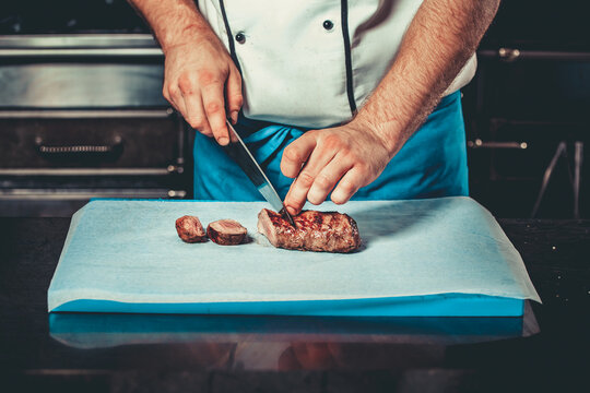Chef in hotel or restaurant kitchen dressed in blue apron cooking, only hands, he is cutting meat or steak with big sharp knife