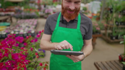 Fototapeta premium Close-up of staff_s hand holding tablet browsing inventory inside local business. Young man wearing green apron using modern technology at Flower Shop