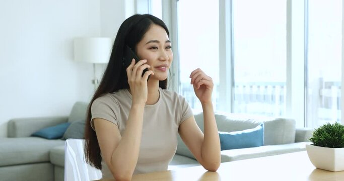 Pretty Asian woman lead pleasant conversation on phone, working in home office, provide information to client, enjoy personal talk to friend, share news speaking on cellphone. Communication use tech