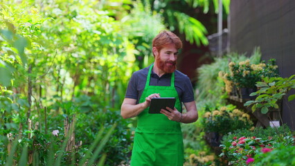 A happy male florist inspecting plant inventory at retail store with tablet. Young man wearing green apron walking toward camera and smiling at camera