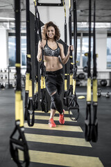 Fototapeta na wymiar Smiling sportive woman with curly hair stands in the gym on the background of the partition. She wears dark sportswear with red sneakers. She looks into the camera and holds TRX straps. Vertical.