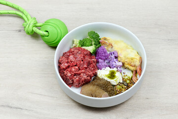 Dog food. Raw meat, chicken, tripe, cauliflower, seeds in white bowl on grey background with green toy ball
