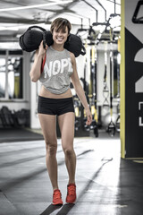 Cute smiling girl stands in the gym on the blurry background. She wears a red top with gray sleeveless, black shorts and red sneakers. She holds a load with right hand on her right shoulder. Vertical.