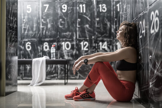 Thoughtful curly woman in red pants and sneakers and black top sits sideways next to the lockers in the dressing-room in the gym. She holds her arms on the knees. Horizontal.