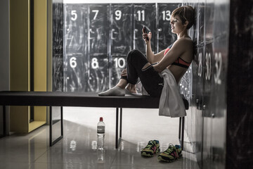 Fototapeta na wymiar Gorgeous smiling girl with a mobile phone sits sideways on the bench in the locker-room in the gym. She wears black pants with white socks and a black-pink top. Horizontal.