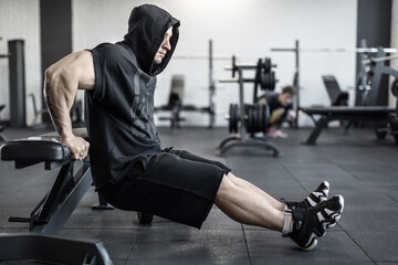 Fototapeta na wymiar Active man with heavy muscles does lifting on the bench in the gym. He wears black hooded sleeveless, black shorts and dark sneakers. Shoot from the side. Horizontal.