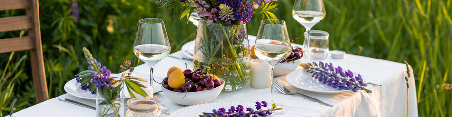 Romantic table decor for a loving couple on the blooming meadow with purple lupines. Two glasses of wine, flowers in a vase, fruits, wooden furniture and picnic basket. Sunset, golden hour. Banner