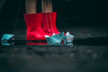 child in red rubber boots holds paper ships in his hands