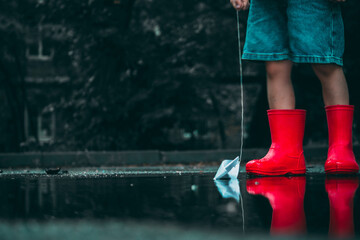 child in red rubber boots holds paper ships in his hands