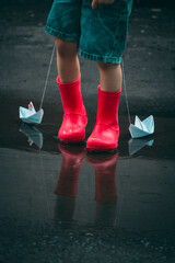 child in red rubber boots holds paper ships in his hands