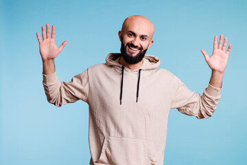 Fototapeta premium Smiling arab man with cheerful facial expression raising hands studio portrait. Young bearded arabian person posing, showing palms and looking at camera with joyful emotions