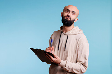 Thoughtful arab man holding clipboard and looking upwards with pensive facial expression. Puzzled...