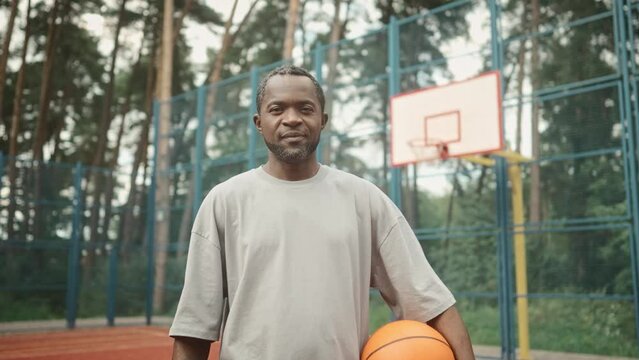 Camera moving to joyful mature man standing on outdoor basketball court. African-American man holds and throws basketball in hands, posing, looking at camera and smiling.