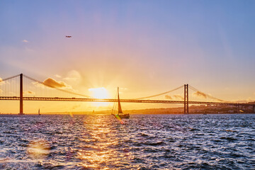 View of 25 de Abril Bridge famous tourist landmark of Lisbon connects Lisboa and Almada on Setubal Peninsula over Tagus river with tourist yacht silhouette at sunset and flying plane. Lisbon, Portugal