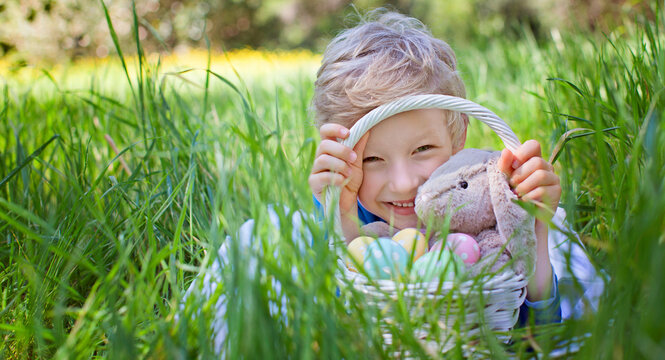 smiling happy boy holding basket with easter eggs and bunny toy after easter egg hunt lying in green grass in the park, copyspace on the left