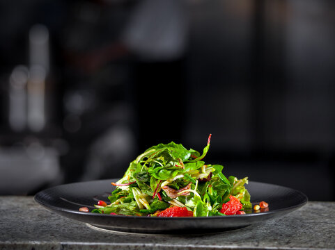 Concept of healthy food. Fresh spring green salad with lettuce, pomegranate and grapefruit, in black plate. Dark interior of modern restaurant kitchen in the background. Ready to eat.