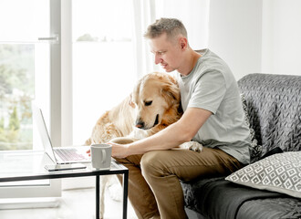 A young man works at a computer with his dog in a bright living room.