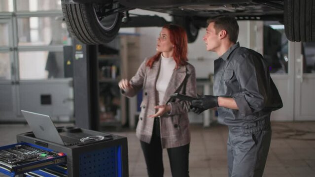smiling woman together with male auto mechanic inspecting motor vehicle on hydraulic ramp