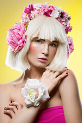 Beautiful young woman in pink dress and blond wig with floral head accessory standing over yellow background. Closeup beauty portrait. Copy space.